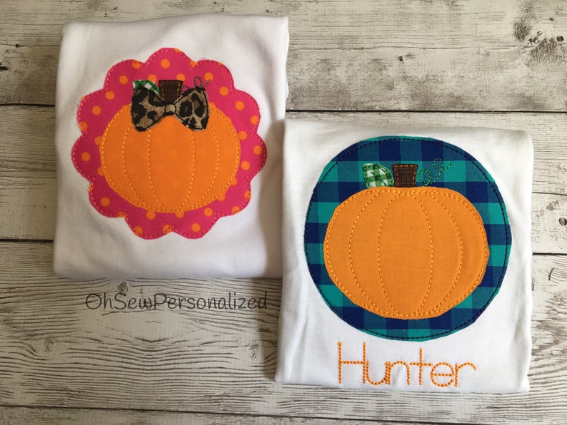 May include: Two white t-shirts with orange pumpkin appliqu&eacute;s. One pumpkin is inside a pink scalloped border with a leopard print bow. The other pumpkin is inside a blue and green plaid circle with the name "Hunter" embroidered below.