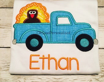 May include: White t-shirt with a blue truck design carrying a turkey. The turkey has a brown body and a colorful fan of feathers. The name "Ethan" is embroidered in orange below the truck.