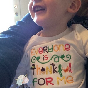 May include: White long-sleeved shirt with the embroidered text "Everyone is thankful for me" in various colors. The design includes a pilgrim hat, acorn, and leaf. The shirt is for a child.