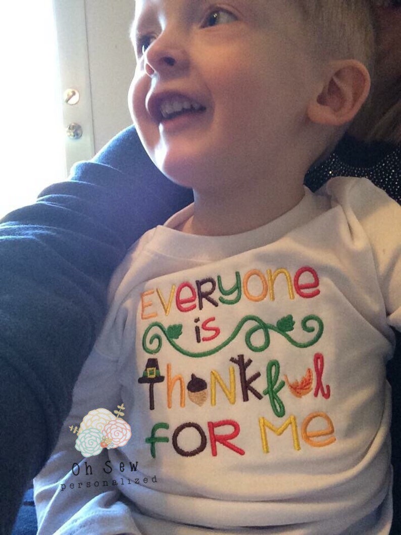 May include: White long-sleeved shirt with the embroidered text "Everyone is thankful for me" in various colors. The design includes a pilgrim hat, acorn, and leaf. The shirt is for a child.