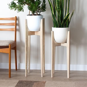 May include: Two white ceramic plant pots with green plants on wooden plant stands. The stands are made of natural wood and have a simple design. A wooden chair is in the background.