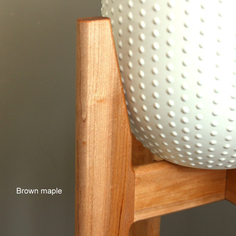 Pode incluir: Um suporte de planta em madeira de bordo castanho claro com um vaso redondo branco texturizado. O suporte tem um design moderno de meados do s&eacute;culo. O texto "Brown maple" &eacute; vis&iacute;vel.