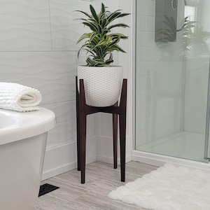 Pode incluir: Um vaso de cer&acirc;mica branca texturizada com uma planta verde e branca est&aacute; em um suporte de planta de madeira marrom escuro. O suporte tem um design moderno de meados do s&eacute;culo com tr&ecirc;s pernas. O suporte da planta est&aacute; em um banheiro.
