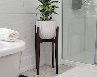 May include: A white textured ceramic planter with a green and white plant sits on a dark brown wooden plant stand. The stand has a mid-century modern design with three legs. The plant stand is in a bathroom setting.