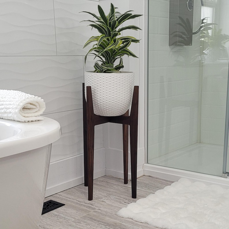 Pode incluir: Um vaso de cer&acirc;mica branca texturizada com uma planta verde e branca est&aacute; em um suporte de planta de madeira marrom escuro. O suporte tem um design moderno de meados do s&eacute;culo com tr&ecirc;s pernas. O suporte da planta est&aacute; em um banheiro.