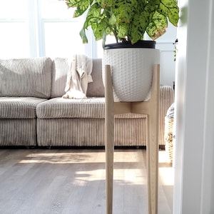Pode incluir: Um vaso de cer&acirc;mica branca texturizada com uma planta verde e branca est&aacute; sobre um suporte de planta de madeira clara. O suporte tem um design minimalista com quatro pernas finas. O fundo inclui um sof&aacute; de veludo cotel&ecirc; e uma janela brilhante.