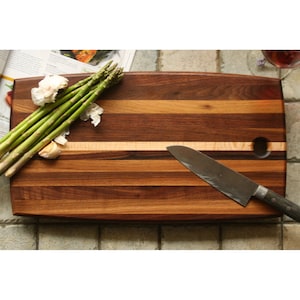 May include: A wooden cutting board with a light brown and dark brown striped pattern. The board has a rounded edge and a hole in the top right corner. A silver knife is lying on the board, and a bunch of asparagus and garlic are on the left side of the board.