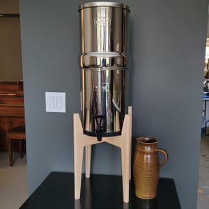 May include: A stainless steel water filter system with a wooden stand and a brown ceramic pitcher. The filter has a spigot and a lid. The pitcher has a handle and a textured surface. The system is on a black surface.
