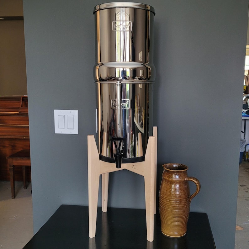 May include: A stainless steel water filter system with a wooden stand and a brown ceramic pitcher. The filter has a spigot and a lid. The pitcher has a handle and a textured surface. The system is on a black surface.