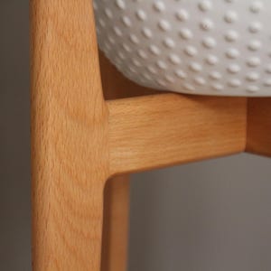 Pode incluir: Close-up de um suporte de planta de madeira clara com uma tigela branca texturizada. O suporte tem um design simples e moderno com linhas limpas e um acabamento natural de gr&atilde;o de madeira. A tigela tem um padr&atilde;o de pequenas sali&ecirc;ncias espa&ccedil;adas.