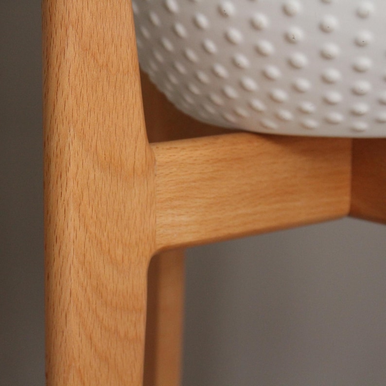 Pode incluir: Close-up de um suporte de planta de madeira clara com uma tigela branca texturizada. O suporte tem um design simples e moderno com linhas limpas e um acabamento natural de gr&atilde;o de madeira. A tigela tem um padr&atilde;o de pequenas sali&ecirc;ncias espa&ccedil;adas.