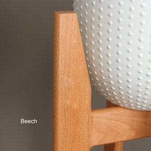 May include: A light-colored wooden plant stand with a white, textured, spherical planter. The stand is made of beech wood and has a simple, modern design. The planter has a pattern of small, raised dots.