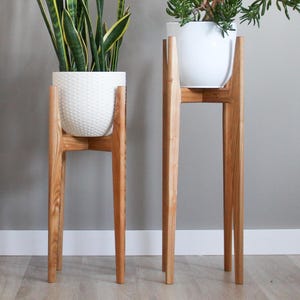Peut inclure: Deux supports de plantes en bois avec des pots en céramique blanche et des plantes. Le support le plus haut est à droite et le plus bas à gauche. Les deux supports ont un design simple avec quatre pieds.