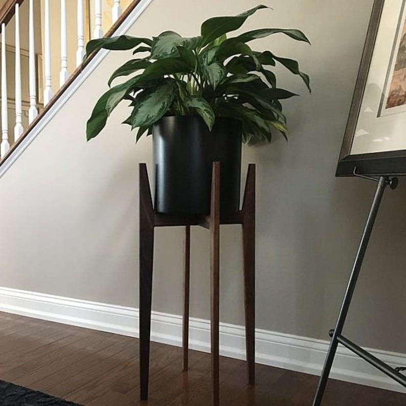 Pode incluir: Uma planta grande e verdejante num vaso preto assenta num suporte de plantas de madeira de estilo moderno de meados do s&eacute;culo. O suporte tem uma base de trip&eacute; com tr&ecirc;s pernas finas e anguladas. O suporte tem cerca de 76 cm de altura.