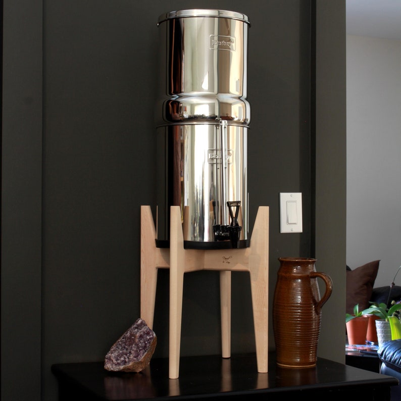 May include: A stainless steel water filter system with a wooden stand. The filter has two stacked cylindrical chambers and a spigot. A brown ceramic pitcher and a purple amethyst crystal are also visible.