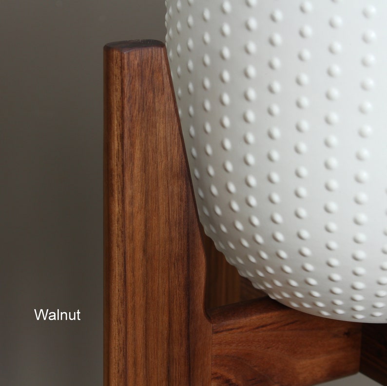 Pode incluir: Um vaso de cer&acirc;mica branca texturizada com um padr&atilde;o pontilhado, suportado por um suporte de madeira de nogueira escura. A palavra "Walnut" est&aacute; impressa no canto inferior esquerdo da imagem. O vaso &eacute; projetado para uso interno ou externo.