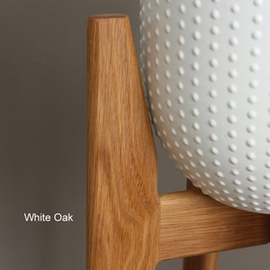 May include: Close-up of a white oak plant stand with a white, textured planter. The wooden stand has a natural finish, showcasing the wood grain. The planter has a pattern of small, raised dots.