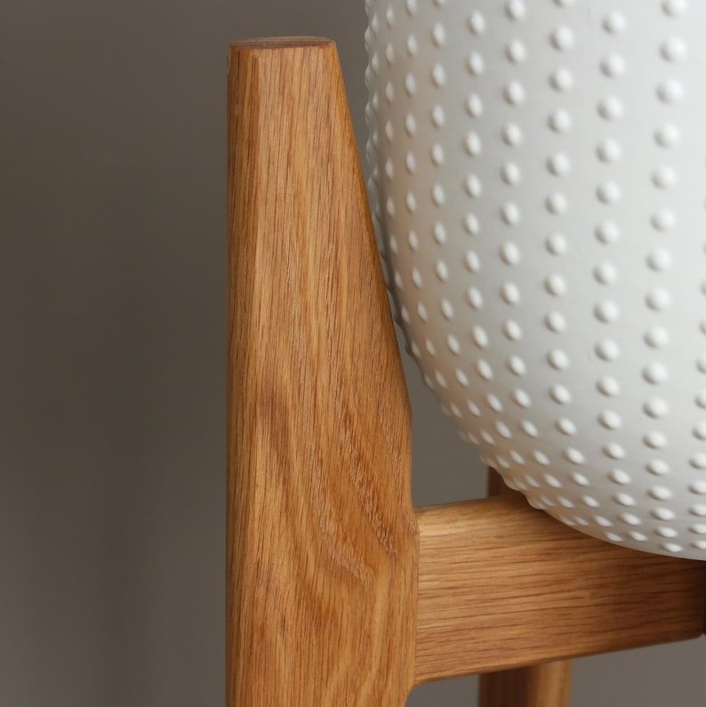 Pode incluir: Uma vista de perto de um suporte de planta de madeira com um vaso branco texturizado. O suporte &eacute; feito de madeira clara e tem um design simples e moderno. O vaso tem um padr&atilde;o de pequenos pontos em relevo.
