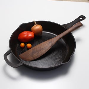 May include: A black cast iron skillet with two side handles and a long handle, containing a dark brown wooden spatula, one red tomato, one yellow onion, and two small yellow cherry tomatoes resting inside the circular cooking surface.