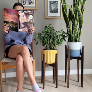 Pode incluir: Uma pessoa sentada numa cadeira de madeira l&ecirc; uma revista intitulada "Affordable Wine". Dois suportes de plantas de madeira seguram um vaso amarelo e um azul e branco com vegeta&ccedil;&atilde;o. Obras de arte emolduradas penduradas na parede cinza.