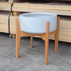 Pode incluir: Um vaso de plantas redondo azul claro com uma superf&iacute;cie texturizada, colocado sobre um suporte de madeira. O suporte tem quatro pernas anguladas e um acabamento em madeira natural. O vaso &eacute; projetado para uso interno ou externo.
