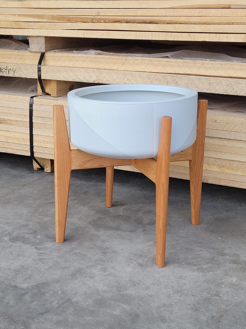Pode incluir: Um vaso de plantas redondo azul claro com uma superf&iacute;cie texturizada, colocado sobre um suporte de madeira. O suporte tem quatro pernas anguladas e um acabamento em madeira natural. O vaso &eacute; projetado para uso interno ou externo.