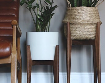 Puede incluir: Dos soportes para plantas de interior. Uno sostiene una maceta de cerámica blanca con una planta verde, el otro una cesta tejida con una planta verde. Ambos están sobre soportes de madera oscura con patas anguladas. Diseño moderno de mediados de siglo.