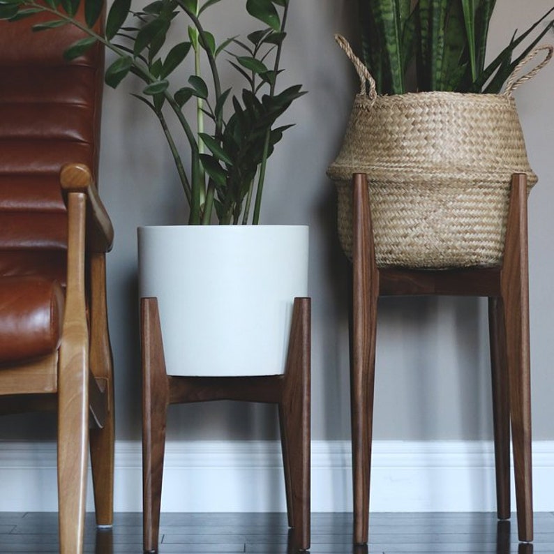 Pode incluir: Dois suportes para plantas de interior. Um deles suporta um vaso de cer&acirc;mica branco com uma planta verde, o outro um cesto tecido com uma planta verde. Ambos est&atilde;o em suportes de madeira escura com pernas anguladas. Design moderno de meados do s&eacute;culo.