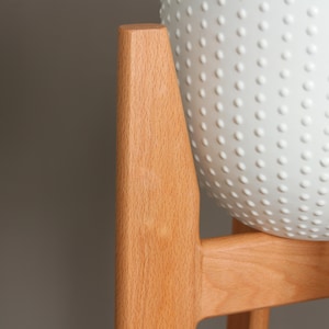 Pode incluir: Um suporte de planta de madeira clara com um vaso branco texturizado. O vaso tem um padr&atilde;o de pequenas sali&ecirc;ncias espa&ccedil;adas uniformemente. O suporte &eacute; feito de madeira clara e tem um design simples e moderno.