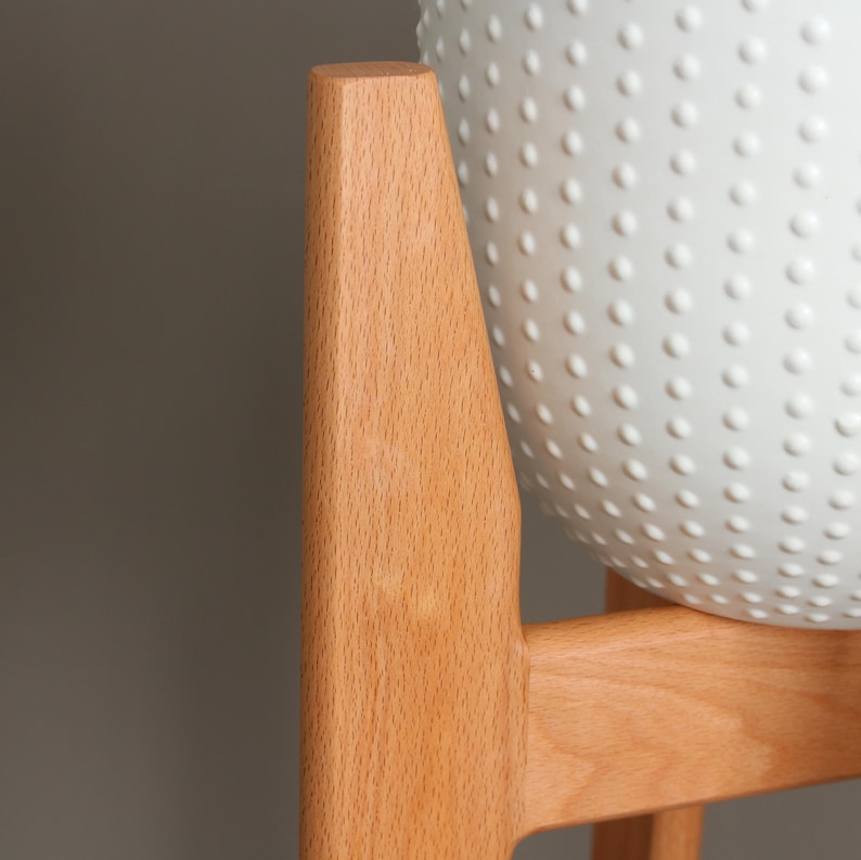 Pode incluir: Um suporte de planta de madeira clara com um vaso branco texturizado. O vaso tem um padr&atilde;o de pequenas sali&ecirc;ncias espa&ccedil;adas uniformemente. O suporte &eacute; feito de madeira clara e tem um design simples e moderno.