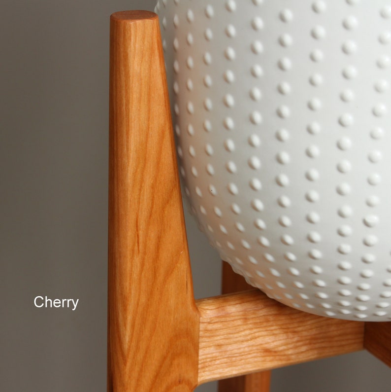 May include: A white ceramic planter with a textured, dotted surface, set on a cherry wood stand. The stand has a mid-century modern design with angled legs and a horizontal support. The word "Cherry" is printed on the image.