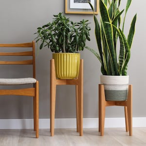 Pode incluir: Uma cadeira de madeira com assento claro &eacute; colocada ao lado de dois suportes de plantas de madeira. Um suporte cont&eacute;m um vaso amarelo canelado com uma planta verde, e o outro um vaso listrado branco e verde com uma planta verde alta.