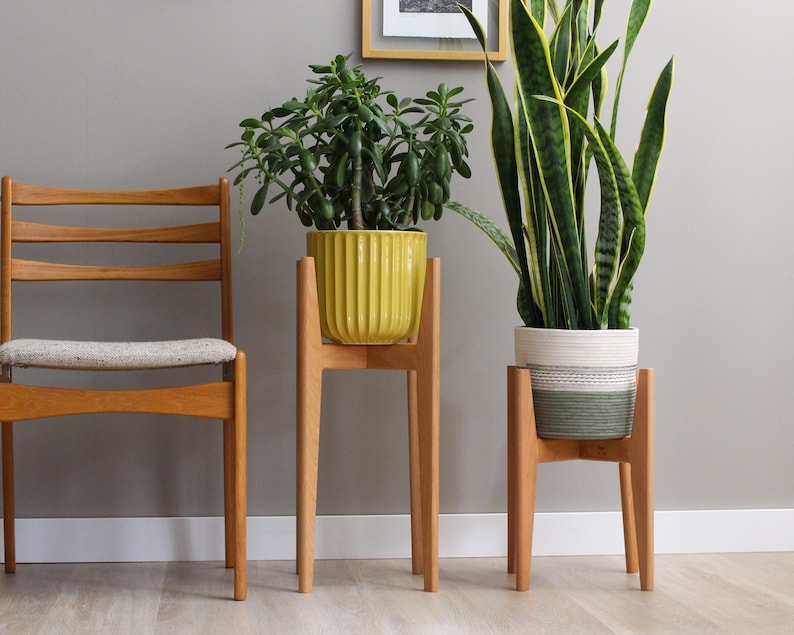 Pode incluir: Uma cadeira de madeira com assento claro &eacute; colocada ao lado de dois suportes de plantas de madeira. Um suporte cont&eacute;m um vaso amarelo canelado com uma planta verde, e o outro um vaso listrado branco e verde com uma planta verde alta.