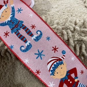 May include: Pink ribbon with a red trim featuring a repeating pattern of elves wearing blue and red outfits with white snowflakes. The elves are holding a holly sprig and have a blue glitter finish.