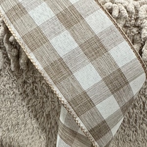 May include: A close-up of a beige and white checkered ribbon. The ribbon has a woven texture and a stitched edge. The ribbon is folded over, revealing the pattern. The background is a textured, light brown surface.
