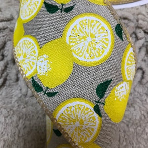 May include: Close-up of a decorative ribbon featuring a repeating pattern of yellow lemons and lemon slices on a tan background. The ribbon's edges are trimmed with a gold-colored stitching, and the design includes green leaves.