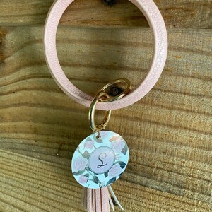 May include: A pink leather key ring with a gold key chain and a round charm with a floral design and the letter "S" in the center. The charm has a pink tassel attached.