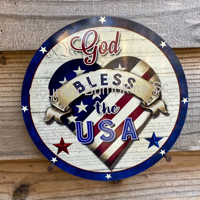 May include: Round patriotic sign with a blue border and stars. The sign features a heart-shaped American flag design with the words "God Bless the USA" in red, white, and blue.