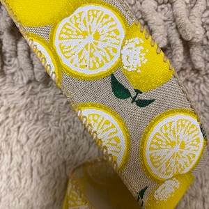 May include: A beige ribbon with a repeating pattern of yellow lemons. The lemons have a white center and are printed on a beige background.