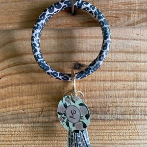 May include: A black and white leopard print key ring with a gold ring and a floral charm with the letter "L" in the center. The charm has a tassel attached.