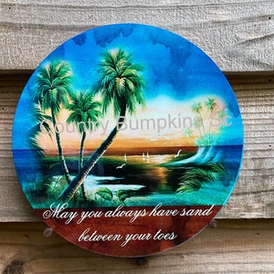 May include: A round, blue and brown ceramic plate with a sunset scene and palm trees. The plate has the text "May you always have sand between your toes" on the bottom.