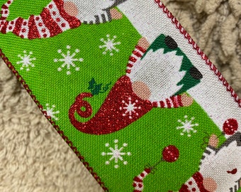 May include: Green and white ribbon with a repeating pattern of gnomes wearing red and white striped stockings. The gnomes are holding red ornaments and are surrounded by white snowflakes.