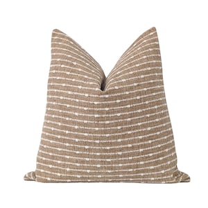 May include: A decorative throw pillow with a neutral color palette. The pillow features a textured, woven fabric in a tan hue, with horizontal rows of small, white dots. The pillow is a square shape, perfect for adding a touch of style to any room.