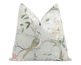 Könnte beinhalten: Ein dekoratives Zierkissen mit hellblauem Hintergrund, das ein Blumen- und Vogelmuster zeigt. Das Design umfasst weiße und rosa Blüten, grüne Blätter und einen Vogel mit blauen, gelben und grünen Federn. Das Kissen hat ein weiches, strukturiertes Aussehen.