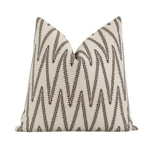May include: A square decorative pillow with a cream-colored background and a repeating pattern of dark brown, jagged lines. The lines create a chevron-like design across the pillow's surface. The pillow is made of a soft fabric.