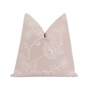 Puede incluir: Un cojín decorativo con fondo rosa claro y un patrón floral blanco. El cojín presenta un diseño detallado de flores, hojas y vides, creando un aspecto clásico y elegante. El color suave y neutro del cojín es adecuado para varios estilos de decoración del hogar.