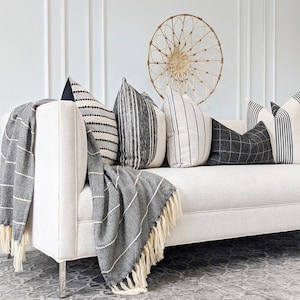 May include: A white couch with black and white striped throw pillows and a black and white striped throw blanket. The couch is in front of a white wall with a woven wall hanging.
