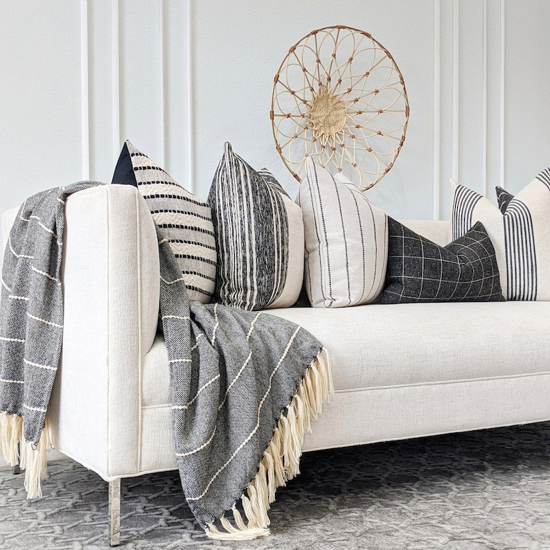 May include: A white couch with black and white striped throw pillows and a black and white striped throw blanket. The couch is in front of a white wall with a woven wall hanging.