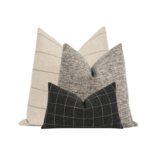 May include: Three decorative throw pillows in a neutral color palette. The largest pillow is a light beige with thin black stripes. The middle pillow is a black and white textured fabric. The smallest pillow is a black and white plaid.