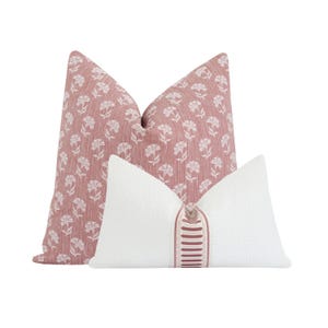 May include: Two decorative throw pillows. One is a dusty rose color with a white floral pattern. The other is white with a textured surface and a vertical stripe of dusty rose and white detail.